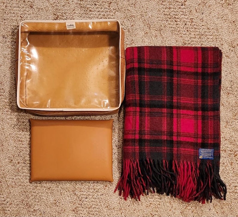 May include: A tan rectangular storage case with a clear plastic top, a solid tan rectangular pouch, and a folded red and black plaid wool blanket with fringe and a Pendleton brand label in the corner. The items are arranged on a textured surface.