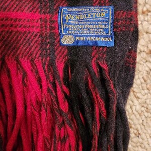 May include: A red and black plaid wool scarf with fringe edges. A blue rectangular label is stitched onto the fabric, reading: Warranted to be a Pendleton, Pendleton Woolen Mills, Portland, Oregon, 100% Virgin Wool, Pure Virgin Wool.