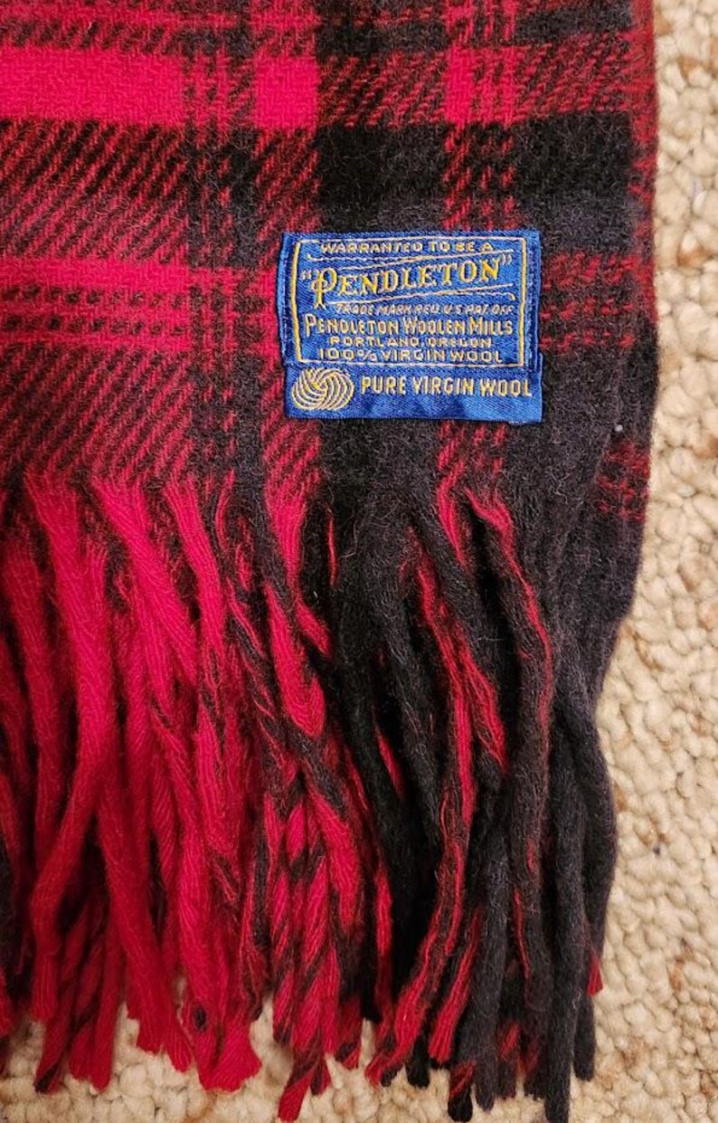 May include: A red and black plaid wool scarf with fringe edges. A blue rectangular label is stitched onto the fabric, reading: Warranted to be a Pendleton, Pendleton Woolen Mills, Portland, Oregon, 100% Virgin Wool, Pure Virgin Wool.