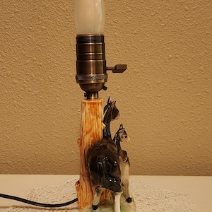 May include: A table lamp featuring a ceramic base with two black and white donkey figures standing beside a brown tree trunk. The lamp has a metallic bronze-colored socket with a turn-knob switch and a single white flame-shaped light bulb.