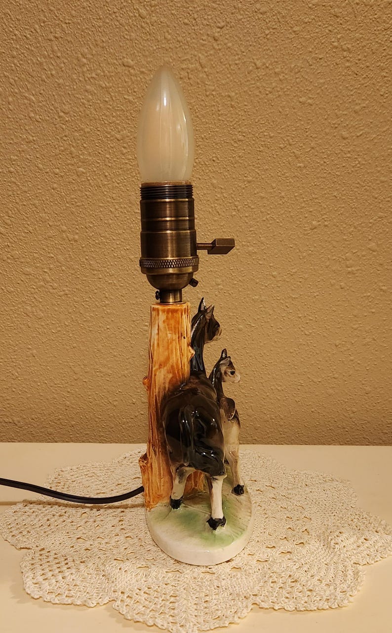 May include: A table lamp featuring a ceramic base with two black and white donkey figures standing beside a brown tree trunk. The lamp has a metallic bronze-colored socket with a turn-knob switch and a single white flame-shaped light bulb.
