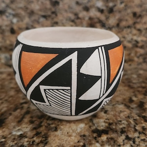 May include: A small, round ceramic bowl with a cream-colored interior and a black, orange, and white geometric design on the exterior. The bowl's design features triangles, lines, and shapes. The bowl is sitting on a brown surface.