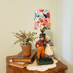 May include: A table lamp with a floral patterned lampshade and a ceramic figurine of a boy and girl. The figurine is painted in bright colors and the lampshade has a white background with pink, blue, and green flowers.