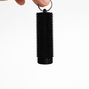 May include: Black cylindrical keychain with a textured, spiky surface. The keychain is held by a hand and features a small metal ring for attaching keys. The design is simple and modern.