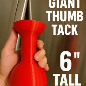 May include: A bright red, oversized thumb tack shaped object with a smooth finish. The text GIANT THUMB TACK 6" TALL is printed in white on the right side. The object features a wide, rounded top and a flared base, standing 6 inches tall.