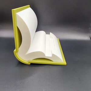 May include: A white book-shaped phone stand with a green border. The stand is made of plastic and has a modern design.