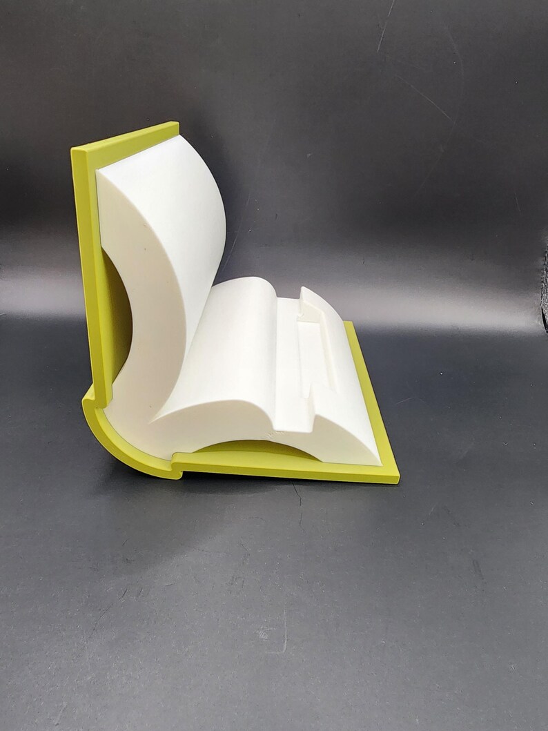May include: A white book-shaped phone stand with a green border. The stand is made of plastic and has a modern design.