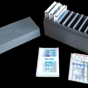 May include: A gray plastic sewing needle storage box with a lid, and a separate open box with dividers. Two packages of sewing machine needles are visible, one labeled "Stretch" and the other "SCHMETZ STRETCH".