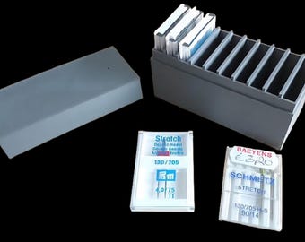 May include: A gray plastic sewing needle storage box with a lid, and a separate open box with dividers. Two packages of sewing machine needles are visible, one labeled "Stretch" and the other "SCHMETZ STRETCH".