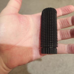 May include: A black, cylindrical object with a textured, spiky surface. The object is held in a hand, showcasing its size and shape. The design appears to be 3D-printed, with a series of small, square-like protrusions covering the entire surface.