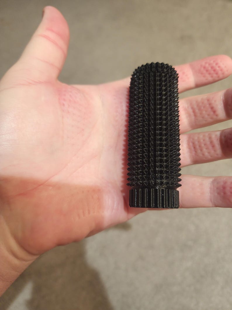 May include: A black, cylindrical object with a textured, spiky surface. The object is held in a hand, showcasing its size and shape. The design appears to be 3D-printed, with a series of small, square-like protrusions covering the entire surface.