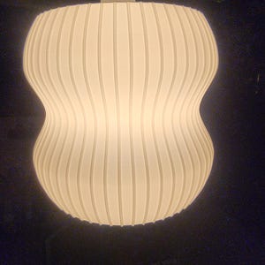 May include: A white, ribbed pendant light fixture with a unique hourglass shape. The light is suspended from a white cord and ceiling mount. The shade has vertical grooves, creating a textured surface. The light is illuminated, casting a warm glow.