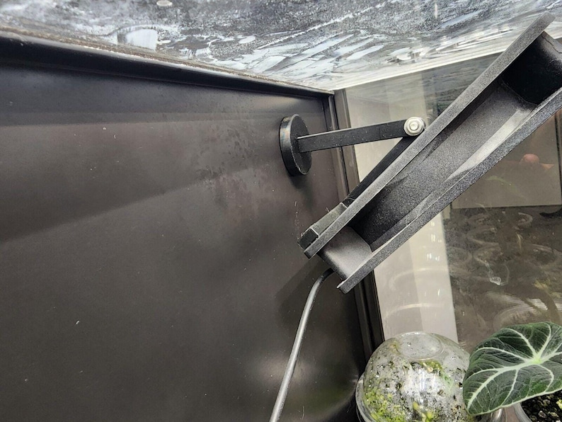 Puede incluir: Un accesorio de luz de metal negro con una base magn&eacute;tica est&aacute; montado en una superficie negra. El accesorio est&aacute; inclinado hacia abajo y conectado a un cable negro.