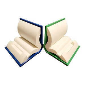 May include: Two book-shaped phone stands, one blue and one green, with a white interior. The stands are open and ready to hold a phone.