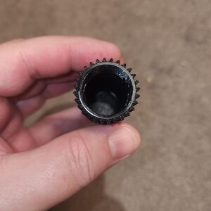 May include: A black, cylindrical object with a textured, ridged exterior and a hollow interior. The top edge features a series of small, pointed protrusions. The object is held in a person's hand.