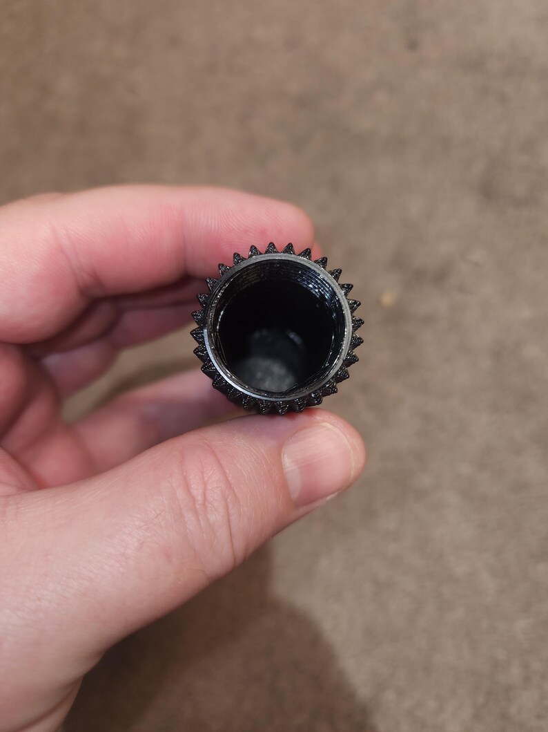 May include: A black, cylindrical object with a textured, ridged exterior and a hollow interior. The top edge features a series of small, pointed protrusions. The object is held in a person's hand.