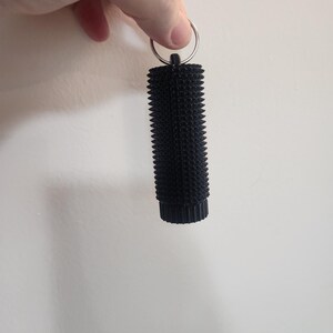 May include: A black, cylindrical keychain with a textured, spiky surface. The keychain is attached to a silver metal ring. The object is held against a plain white background.