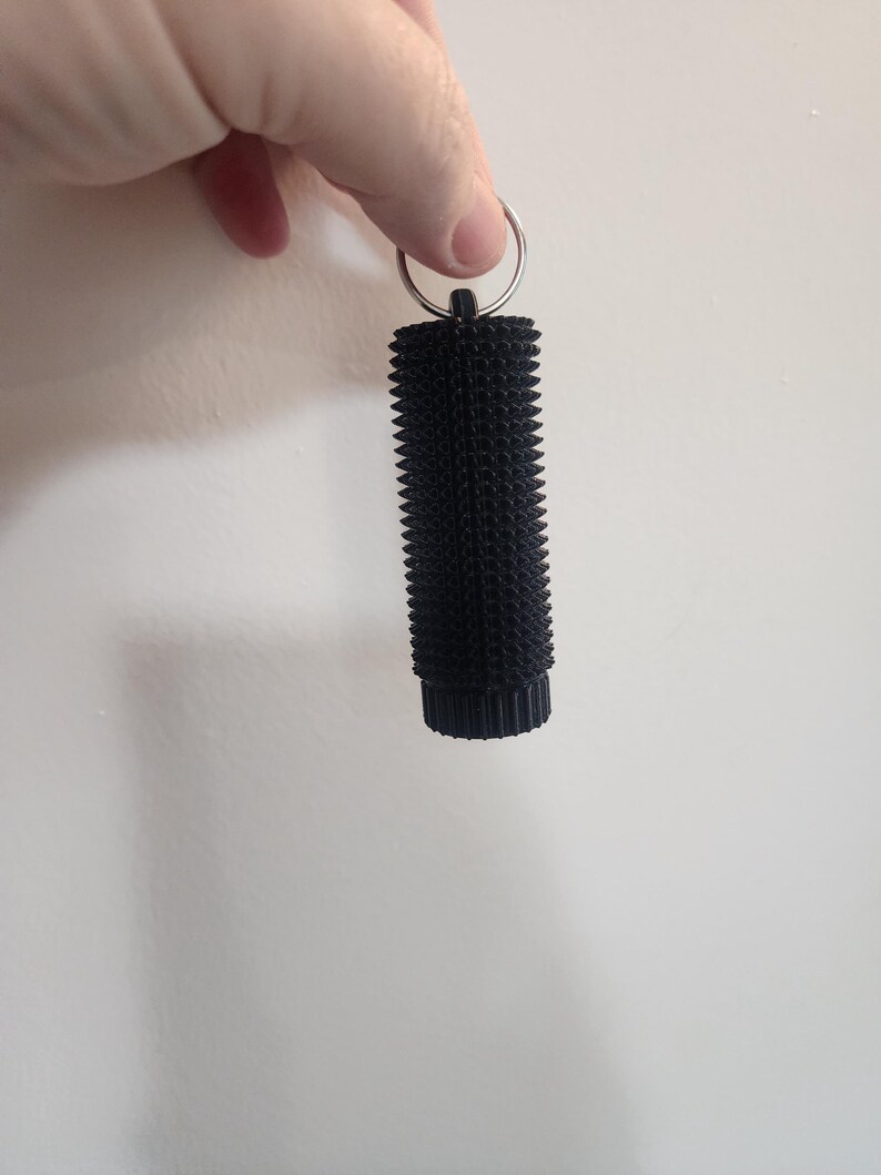 May include: A black, cylindrical keychain with a textured, spiky surface. The keychain is attached to a silver metal ring. The object is held against a plain white background.