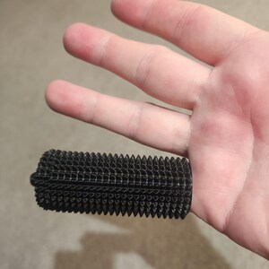May include: A black, rectangular object with a textured, spiky surface. The object is held in a hand, showcasing its detailed design. The object appears to be a grip or handle, possibly for a tool or device.