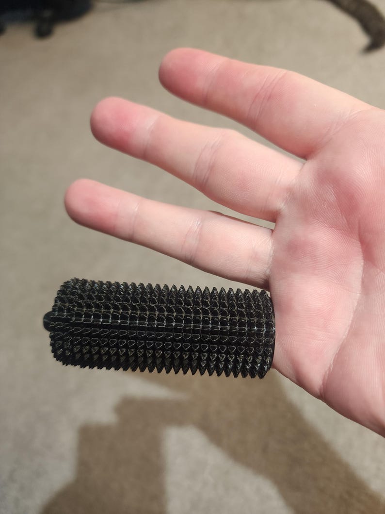 May include: A black, rectangular object with a textured, spiky surface. The object is held in a hand, showcasing its detailed design. The object appears to be a grip or handle, possibly for a tool or device.