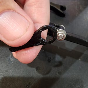Puede incluir: Una pieza de pl&aacute;stico negro con un agujero hexagonal, unida a un brazo de pl&aacute;stico negro con una tuerca plateada.