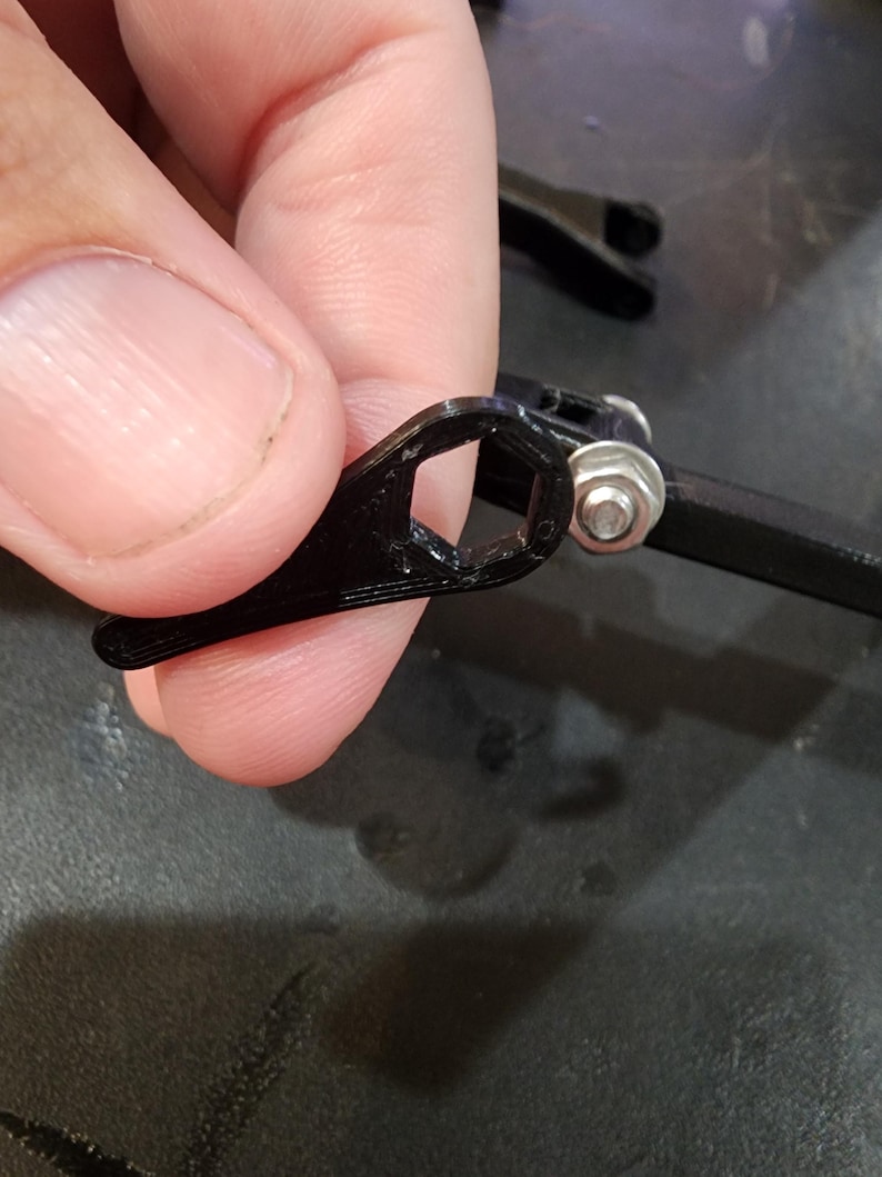 Puede incluir: Una pieza de pl&aacute;stico negro con un agujero hexagonal, unida a un brazo de pl&aacute;stico negro con una tuerca plateada.
