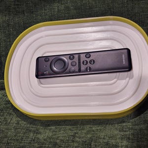 May include: A black Samsung TV remote control rests on a white and yellow oval-shaped tray. The remote has multiple buttons and a circular control pad. The tray has a layered design.