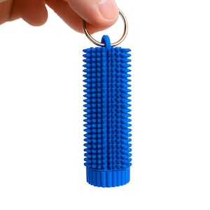 May include: A blue cylindrical pet hair remover with a textured surface and a metal keyring. The tool is designed to remove pet hair from clothing and other surfaces. The item is held by a hand against a white background.