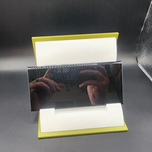 May include: A black smartphone with a reflective screen is displayed on a yellow stand. The screen reflects a hand holding the phone.