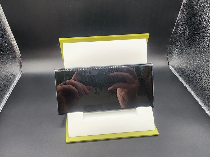 May include: A black smartphone with a reflective screen is displayed on a yellow stand. The screen reflects a hand holding the phone.
