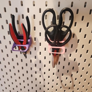 May include: A white pegboard with two sets of tools. On the left, black and red pliers rest in a purple holder. On the right, black scissors are held by a pink holder. The pegboard has evenly spaced holes.