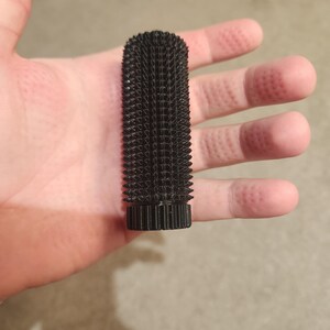 May include: A black, cylindrical object with a textured surface covered in small, pointed protrusions. The object has a ridged base and a rough, spiky appearance. The item is held in a hand.