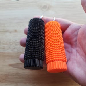 May include: Two textured, cylindrical objects. One is black, the other is bright orange. Both have a ridged base and a textured surface. Each has a small metal loop at the top. The objects are held in a hand.