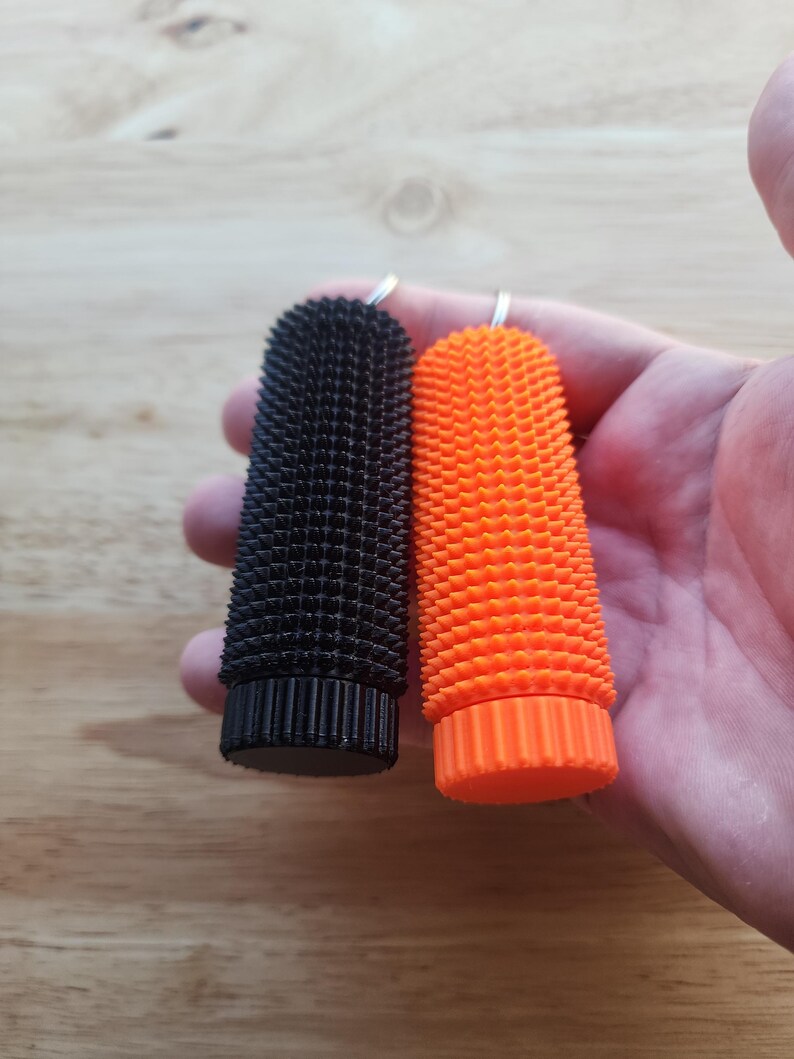 May include: Two textured, cylindrical objects. One is black, the other is bright orange. Both have a ridged base and a textured surface. Each has a small metal loop at the top. The objects are held in a hand.