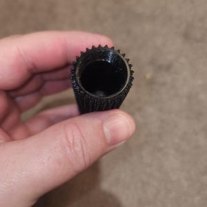 May include: A black, cylindrical object with a textured surface and a toothed rim. The interior is hollow and dark. The object is held in a person's hand.