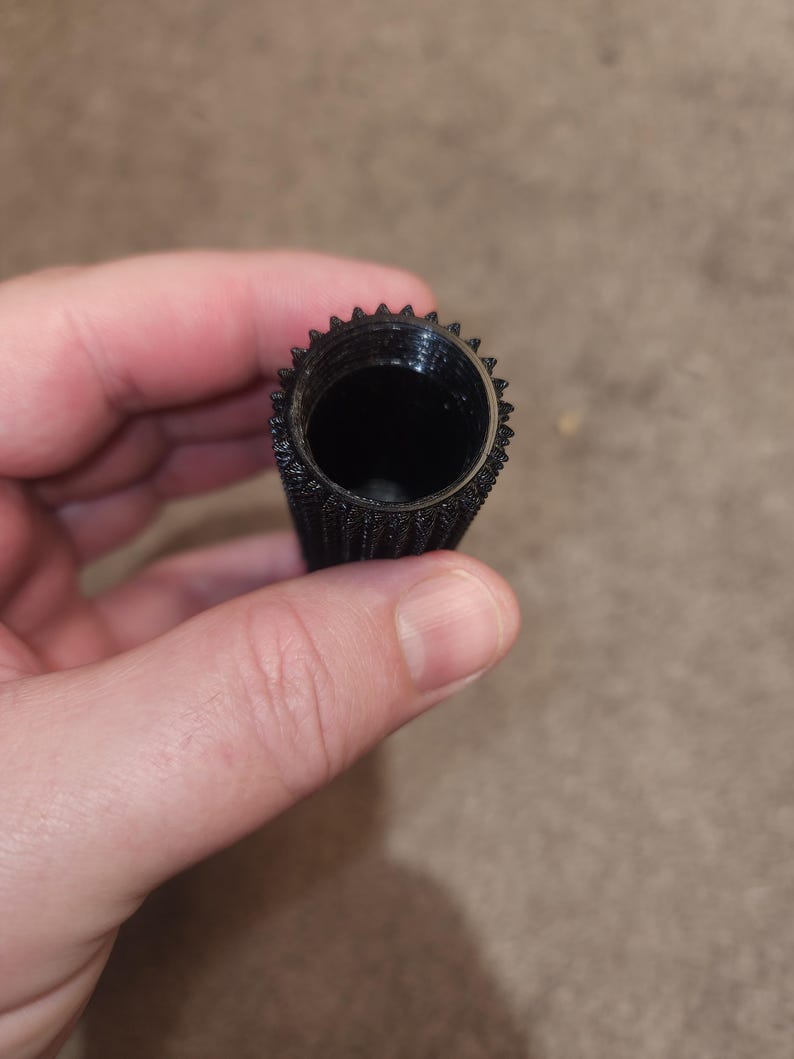 May include: A black, cylindrical object with a textured surface and a toothed rim. The interior is hollow and dark. The object is held in a person's hand.