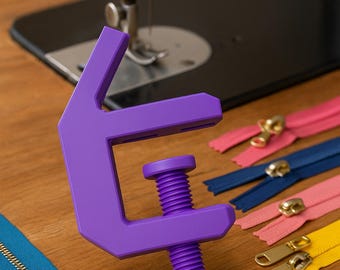 May include: A purple zipper pull tool with a screw mechanism is displayed on a wooden surface, alongside colorful zippers in pink, yellow, and blue. The text "MAKE ZIPPER PULLS LIKE A PRO" is at the top of the image.