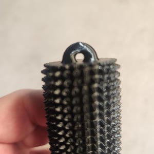 May include: A black, cylindrical rasp tool with a textured surface and a loop at the top. The rasp has a series of small, pointed teeth covering its surface. The tool is designed for shaping and smoothing wood or other materials.