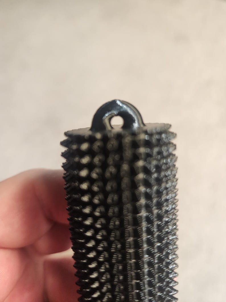 May include: A black, cylindrical rasp tool with a textured surface and a loop at the top. The rasp has a series of small, pointed teeth covering its surface. The tool is designed for shaping and smoothing wood or other materials.