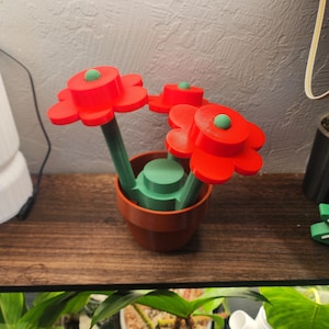 May include: A decorative flower arrangement featuring three red, stylized flowers with green centers and stems. The flowers are in a brown pot with a green base. The arrangement is placed on a wooden surface.