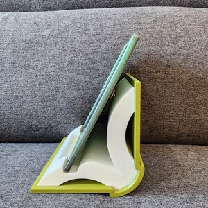 May include: A green tablet stand made of plastic, shaped like an open book. The stand is designed to hold a tablet in an upright position, allowing for easy viewing.