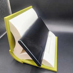 May include: A green and white phone stand with a black phone resting in it. The stand is made of two pieces that are joined together to form a curved shape.