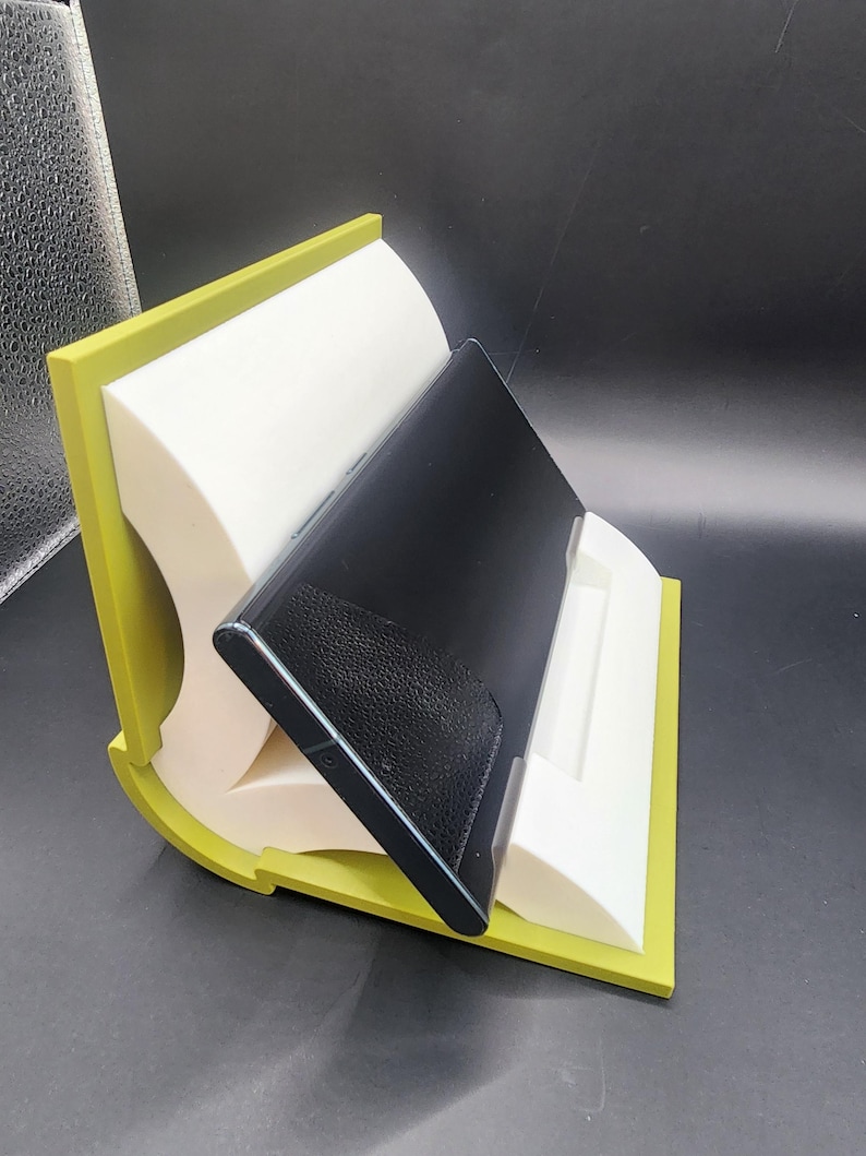 May include: A green and white phone stand with a black phone resting in it. The stand is made of two pieces that are joined together to form a curved shape.