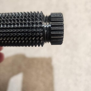 May include: Close-up of a black, textured cylindrical object with a ridged end. The object has a detailed, patterned surface and a threaded section. The lighting highlights the intricate design and the dark color of the material.