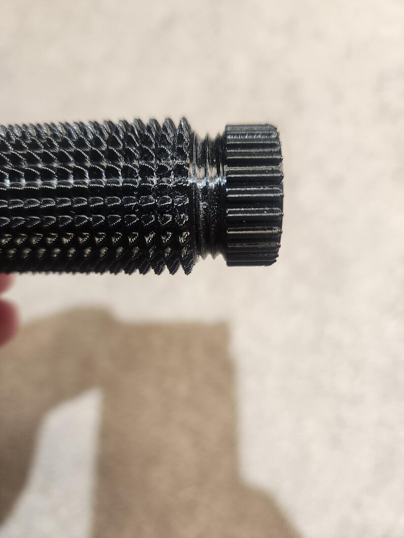 May include: Close-up of a black, textured cylindrical object with a ridged end. The object has a detailed, patterned surface and a threaded section. The lighting highlights the intricate design and the dark color of the material.