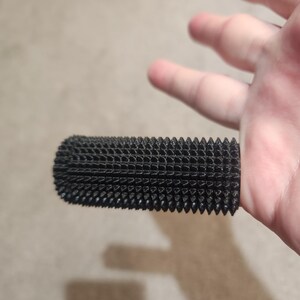 May include: A black, cylindrical object with a textured, grid-like surface and numerous small, pointed protrusions. The object appears to be made of a porous material and is held by a hand.