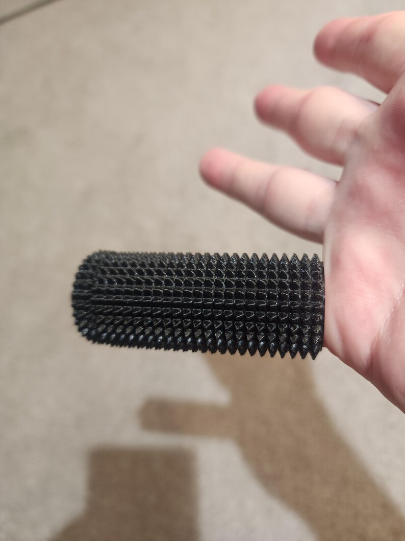 May include: A black, cylindrical object with a textured, grid-like surface and numerous small, pointed protrusions. The object appears to be made of a porous material and is held by a hand.