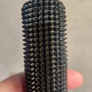 May include: A black, cylindrical object with a textured surface covered in small, rounded protrusions. The object appears to be made of a dark material and has a slightly tapered shape. The design is symmetrical and repetitive.