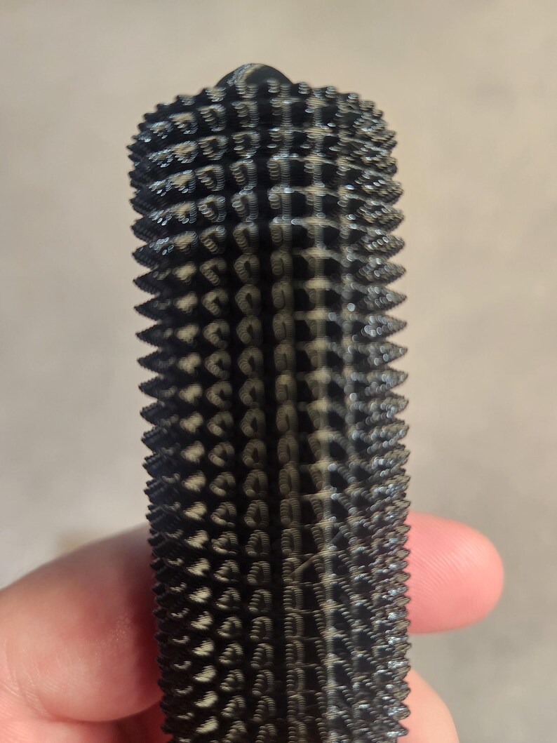May include: A black, cylindrical object with a textured surface covered in small, rounded protrusions. The object appears to be made of a dark material and has a slightly tapered shape. The design is symmetrical and repetitive.