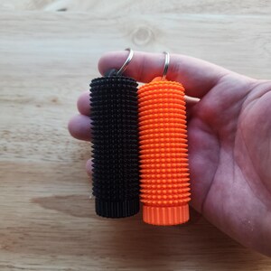May include: Two textured keychains, one black and one orange, are held up against a wooden background. The keychains have a cylindrical shape with a spiky surface. Each keychain has a silver keyring attached to the top.